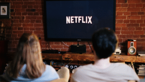 15 Comforting Netflix Movies .Person sitting on a couch under a blanket with popcorn, watching Netflix on a TV in a warm, softly lit living room